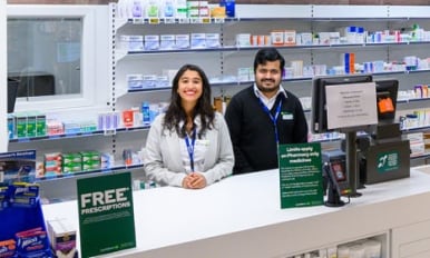 A pair of helpful pharmacy staff look at the viewer with friendly expressions.